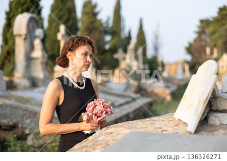 Woman in black with flowers standing by grave in cemetery Woman in black with flowers standing by grave in cemetery 136326271
