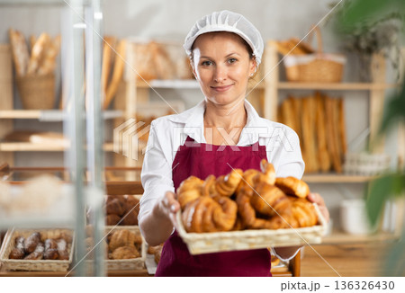 Female baker in white uniform stands behind counter in small private bakery offering hot croissants 136326430