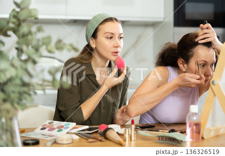 Two female friends applying makeup by vanity mirror at home 136326519