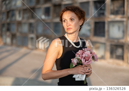 Upset woman in black dress, who lost loved one, brings flowers to grave 136326526