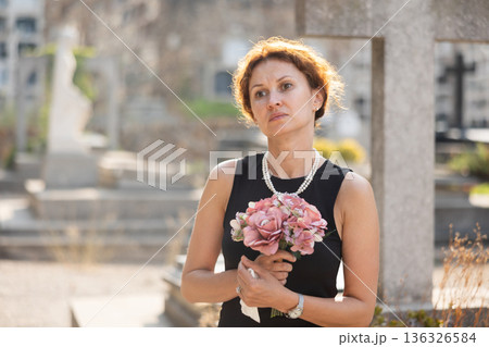 Grieving woman in black with flowers standing in cemetery Grieving woman in black with flowers standing in cemetery 136326584