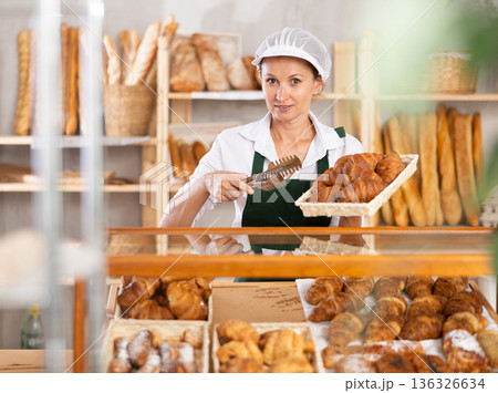 Woman employee puts croissants in window, arranges display of goods at bakery. Woman employee puts croissants in window, arranges display of goods at bakery. 136326634