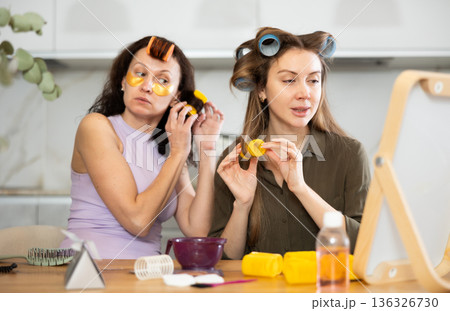 Two adult besties applying hair curlers in front of tabletop mirror Two adult besties applying hair curlers in front of tabletop mirror 136326730