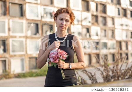 Sad woman in black dress with bouquet of flowers and handkerchief in hands, stands in cemetery 136326881
