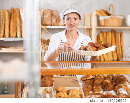 Positive female bakery seller with tongs in his hands offers to buy fresh croissants Positive female bakery seller with tongs in his hands offers to buy fresh croissants 136326961