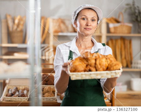 Female baker in white uniform stands behind counter in small private bakery offering hot croissants 136327100