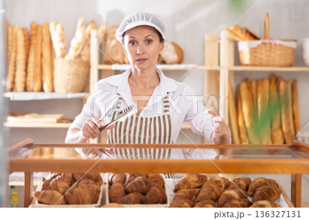 Positive female bakery seller with tongs in his hands offers to buy fresh croissants Positive female bakery seller with tongs in his hands offers to buy fresh croissants 136327131