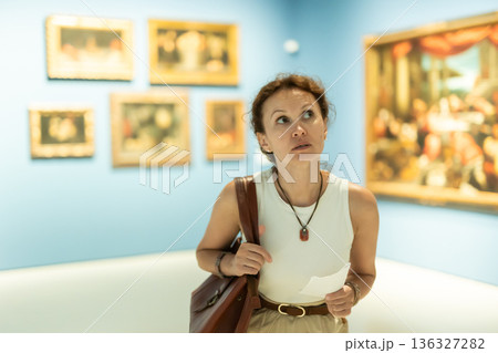 Brunette woman visitor holding guide list standing near pictures in museum of arts Brunette woman visitor holding guide list standing near pictures in museum of arts 136327282