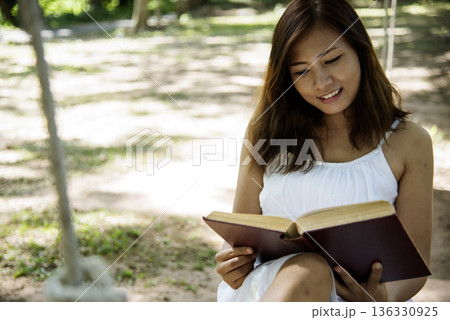Asian woman holding book reading magazine at green park in natural garden. Young woman relaxation read open book self study. University women smiling with happiness learning. Women sit in green park Asian woman holding book reading magazine at green park in natural garden. Young woman relaxation read open book self study. University women smiling with happiness learning. Women sit in green park 136330925