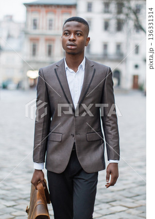 A young Black man in a suit walks down a cobblestone street carrying a leather briefcase, looking thoughtfully into the distance A young Black man in a suit walks down a cobblestone street carrying a leather briefcase, looking thoughtfully into the distance 136331314