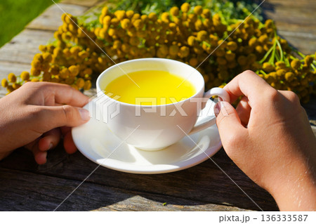 A hot tansy infusion, known for its antispasmodic and choleretic effects, is served in an elegant white cup against a natural wooden background. 136333587