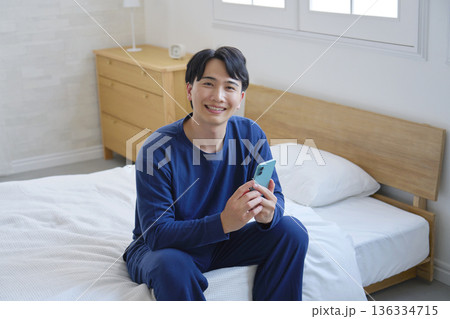 ベッドに座ってスマホを持つ20代男性　カメラ目線　ポートレート 136334715