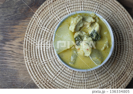 Homemade green curry featuring white gourd and fish balls is served in a gray ceramic bowl set on a circular woven placemat. 136334805