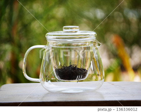 Clear glass teapot with loose black tea leaves inside, placed on wooden table with natural green bokeh background. Minimal tea brewing concept, calm lifestyle, healthy drink, clean design, relaxation 136336012