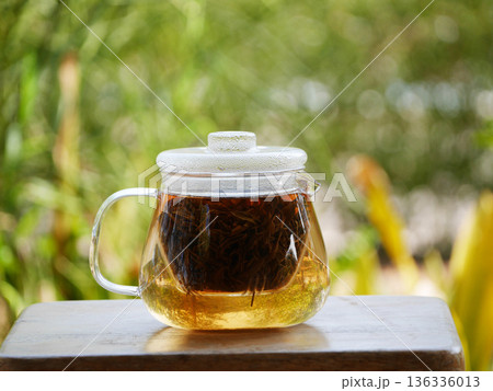 glass teapot brewing loose black tea with hot water, golden tea color visible inside. Minimal tea preparation scene on wooden table with natural green bokeh background, calm lifestyle, healthy drink glass teapot brewing loose black tea with hot water, golden tea color visible inside. Minimal tea preparation scene on wooden table with natural green bokeh background, calm lifestyle, healthy drink 136336013
