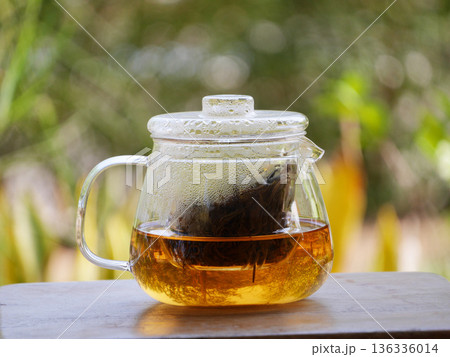 glass teapot brewing loose black tea with hot water, golden tea color visible inside. Minimal tea preparation scene on wooden table with natural green bokeh background, calm lifestyle, healthy drink 136336014