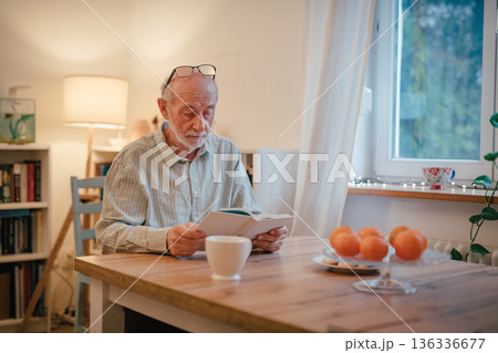 Older man reading book i kitchen. 136336677