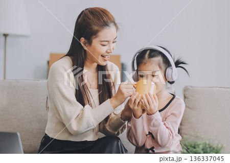 A cheerful Asian mother helps her young daughter do homework and color a book at home, while the father works on a laptop in the background. A cozy, bright, and modern family lifestyle. 136337045