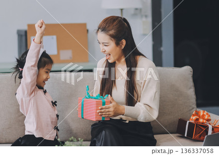A cheerful Asian mother helps her young daughter do homework and color a book at home, while the father works on a laptop in the background. A cozy, bright, and modern family lifestyle. 136337058