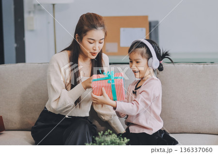 A cheerful Asian mother helps her young daughter do homework and color a book at home, while the father works on a laptop in the background. A cozy, bright, and modern family lifestyle. 136337060