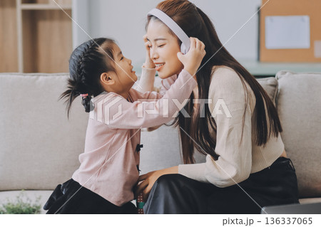 A cheerful Asian mother helps her young daughter do homework and color a book at home, while the father works on a laptop in the background. A cozy, bright, and modern family lifestyle. 136337065