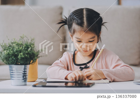 A cheerful Asian mother helps her young daughter do homework and color a book at home, while the father works on a laptop in the background. A cozy, bright, and modern family lifestyle. 136337080