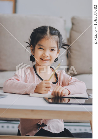 A cheerful Asian mother helps her young daughter do homework and color a book at home, while the father works on a laptop in the background. A cozy, bright, and modern family lifestyle. 136337082