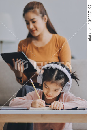 A cheerful Asian mother helps her young daughter do homework and color a book at home, while the father works on a laptop in the background. A cozy, bright, and modern family lifestyle. A cheerful Asian mother helps her young daughter do homework and color a book at home, while the father works on a laptop in the background. A cozy, bright, and modern family lifestyle. 136337107