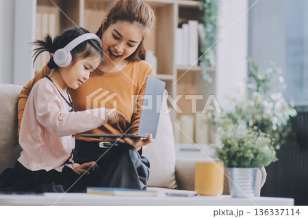 A cheerful Asian mother helps her young daughter do homework and color a book at home, while the father works on a laptop in the background. A cozy, bright, and modern family lifestyle. 136337114