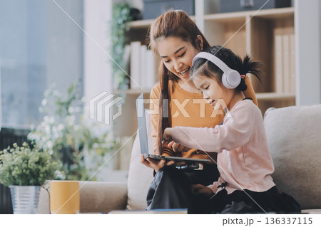 A cheerful Asian mother helps her young daughter do homework and color a book at home, while the father works on a laptop in the background. A cozy, bright, and modern family lifestyle. 136337115