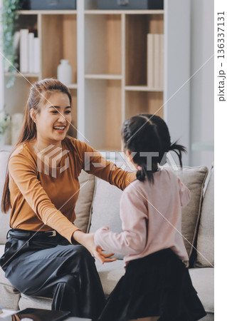 A cheerful Asian mother helps her young daughter do homework and color a book at home, while the father works on a laptop in the background. A cozy, bright, and modern family lifestyle. 136337131