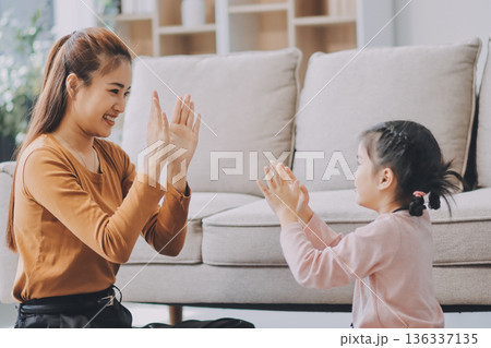 A cheerful Asian mother helps her young daughter do homework and color a book at home, while the father works on a laptop in the background. A cozy, bright, and modern family lifestyle. A cheerful Asian mother helps her young daughter do homework and color a book at home, while the father works on a laptop in the background. A cozy, bright, and modern family lifestyle. 136337135