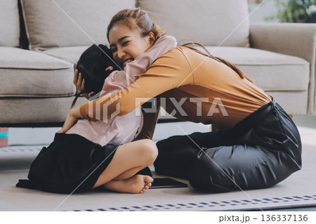 A cheerful Asian mother helps her young daughter do homework and color a book at home, while the father works on a laptop in the background. A cozy, bright, and modern family lifestyle. 136337136
