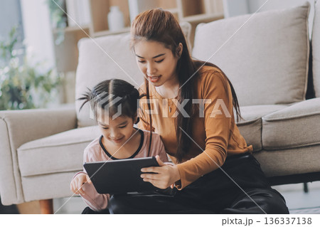 A cheerful Asian mother helps her young daughter do homework and color a book at home, while the father works on a laptop in the background. A cozy, bright, and modern family lifestyle. A cheerful Asian mother helps her young daughter do homework and color a book at home, while the father works on a laptop in the background. A cozy, bright, and modern family lifestyle. 136337138