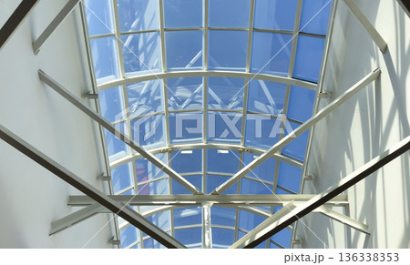Modern glass ceiling with metal structural beams viewed from below against a clear blue sky. 136338353