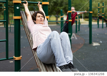 Mature woman goes in for sports in city park on sports ground - doing exercises to pull his legs to chest Mature woman goes in for sports in city park on sports ground - doing exercises to pull his legs to chest 136341208