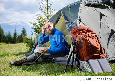 Man sits in tourist tent's entrance, studying map with thoughtful expression. Beside him orange backpack, equipped with tripod and solar panel, set against scenic background of trees and mountains. Man sits in tourist tent's entrance, studying map with thoughtful expression. Beside him orange backpack, equipped with tripod and solar panel, set against scenic background of trees and mountains. 136341678