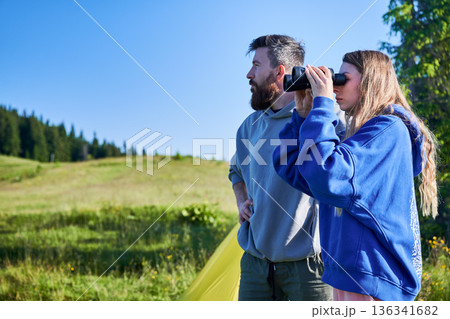 Man and woman stand in grassy field under clear blue sky. Woman, in blue hoodie, looks through binoculars while man, in gray hoodie, stands beside her. Pine trees in background. Man and woman stand in grassy field under clear blue sky. Woman, in blue hoodie, looks through binoculars while man, in gray hoodie, stands beside her. Pine trees in background. 136341682