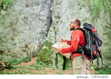 Bearded tourist man studies map while stands in front of large rock in dense forest. Traveler with grey backpack and glasses, planning outdoor adventure, hiking or climbing route. 136341683