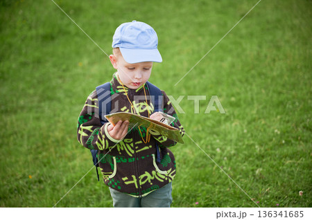 Young boy hiking in grassy field, attentively studying map and compass outdoors. With backpack on shoulders, tourist focused on navigating or planning his route in the mountains. Young boy hiking in grassy field, attentively studying map and compass outdoors. With backpack on shoulders, tourist focused on navigating or planning his route in the mountains. 136341685