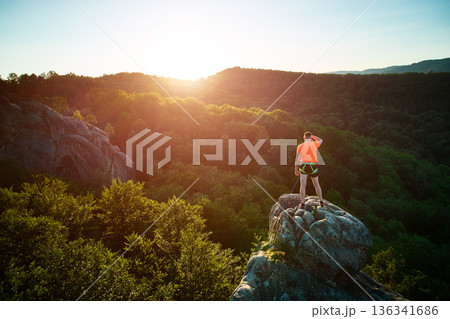 Aerial view of climber standing atop rock formation at Dovbush Rocks in Carpathian mountains, Ukraine. Sun sets, casting warm glow over lush, green forest, distant hills, creating breathtaking view. 136341686