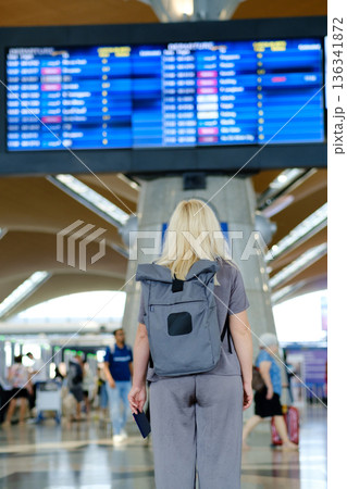 Traveler waiting at the airport terminal looking at the flight information board displaying departure and arrival times in a busy travel environment Traveler waiting at the airport terminal looking at the flight information board displaying departure and arrival times in a busy travel environment 136341872