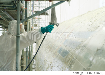 Worker in protective gear spray painting a large industrial tank at a construction site during daylight hours 136341879