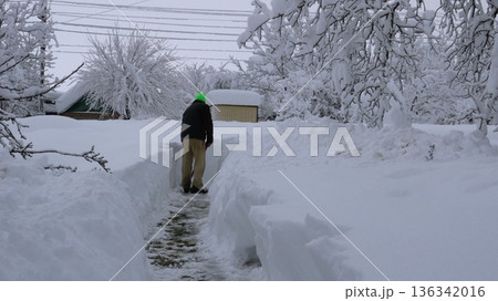 A man in warm clothing, seen from behind, clears a path through high snowdrifts on a residential property leading to a roadway in a rural area. Clearing a path leading from a house to a road  136342016