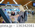 Woman using a touchscreen information kiosk at an airport terminal while travelers wait in the busy background during the daytime 136342081