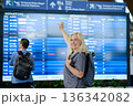 Woman with blonde hair smiles and points at departure board while standing at busy airport terminal in the afternoon, preparing for her upcoming flight departing soon 136342082