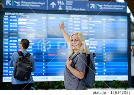 Woman with blonde hair smiles and points at departure board while standing at busy airport terminal in the afternoon, preparing for her upcoming flight departing soon Woman with blonde hair smiles and points at departure board while standing at busy airport terminal in the afternoon, preparing for her upcoming flight departing soon 136342082