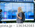 Woman with blonde hair stands near departure board at an airport, ready for an upcoming flight while travellling with a backpack on a sunny day 136342083