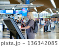 Woman checks in at an airport self-service kiosk while traveling, with busy terminal in the background during afternoon hours 136342085
