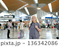 Woman standing in a busy international airport terminal with people walking around and modern architecture in the background during daytime travel hours 136342086